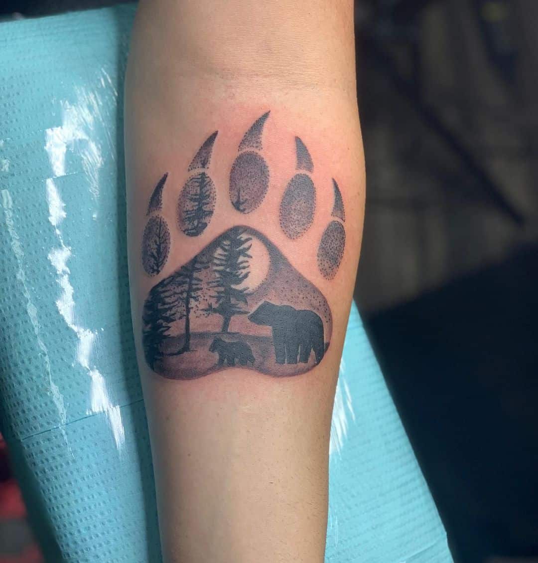 Tattoo of a bear paw with trees and a bear silhouette, elegantly inked on the arm
