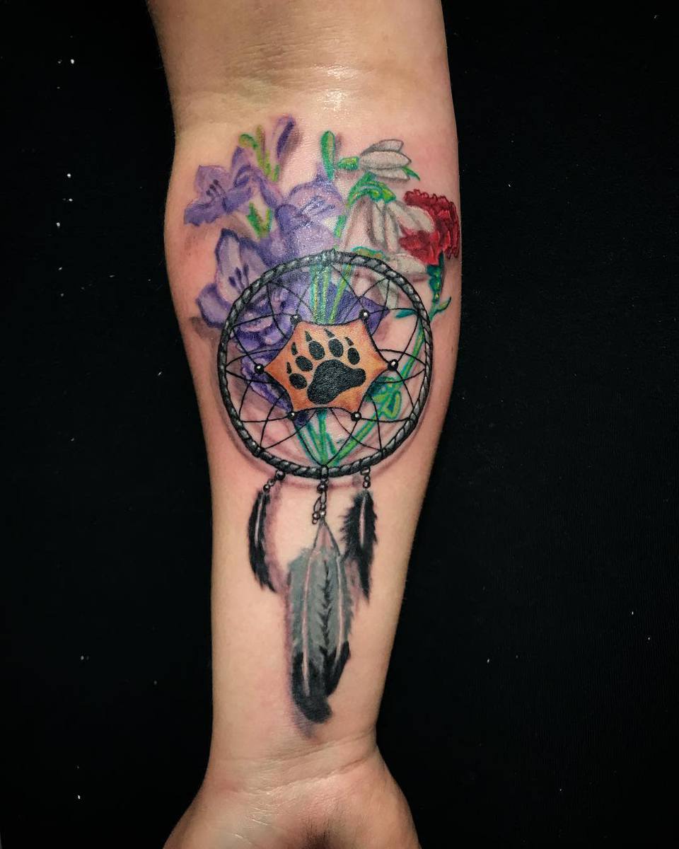 Forearm tattoo of a dreamcatcher with flowers, a bear paw print, and three feathers hanging down