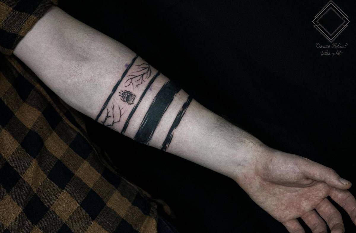 Forearm tattoo with black stripes, tree branches, and a small anatomical heart design, featuring a subtle bear paw for added intrigue