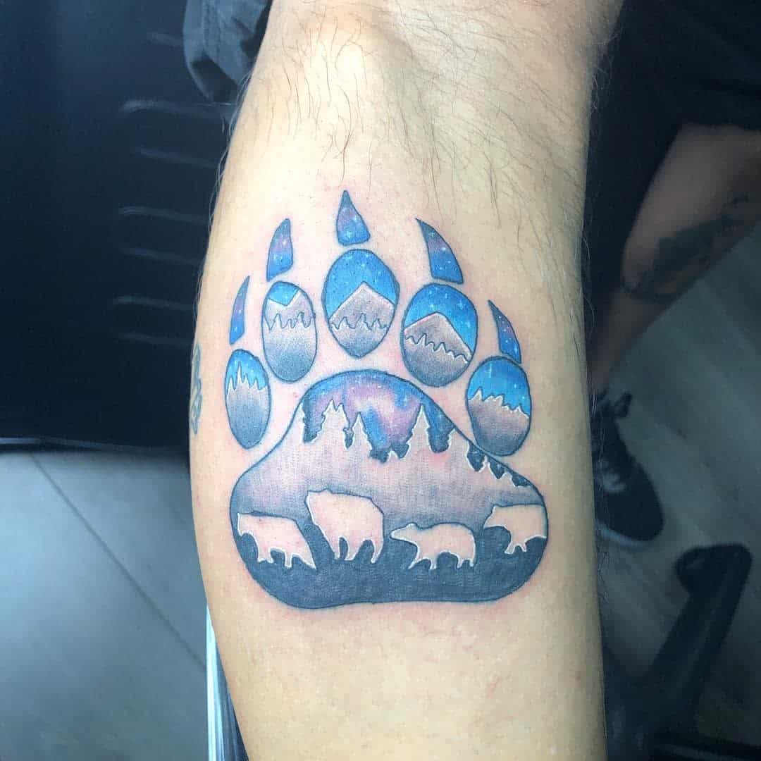 Tattoo of a bear paw with mountain, forest, and bear silhouettes intricately designed within it on a person's arm