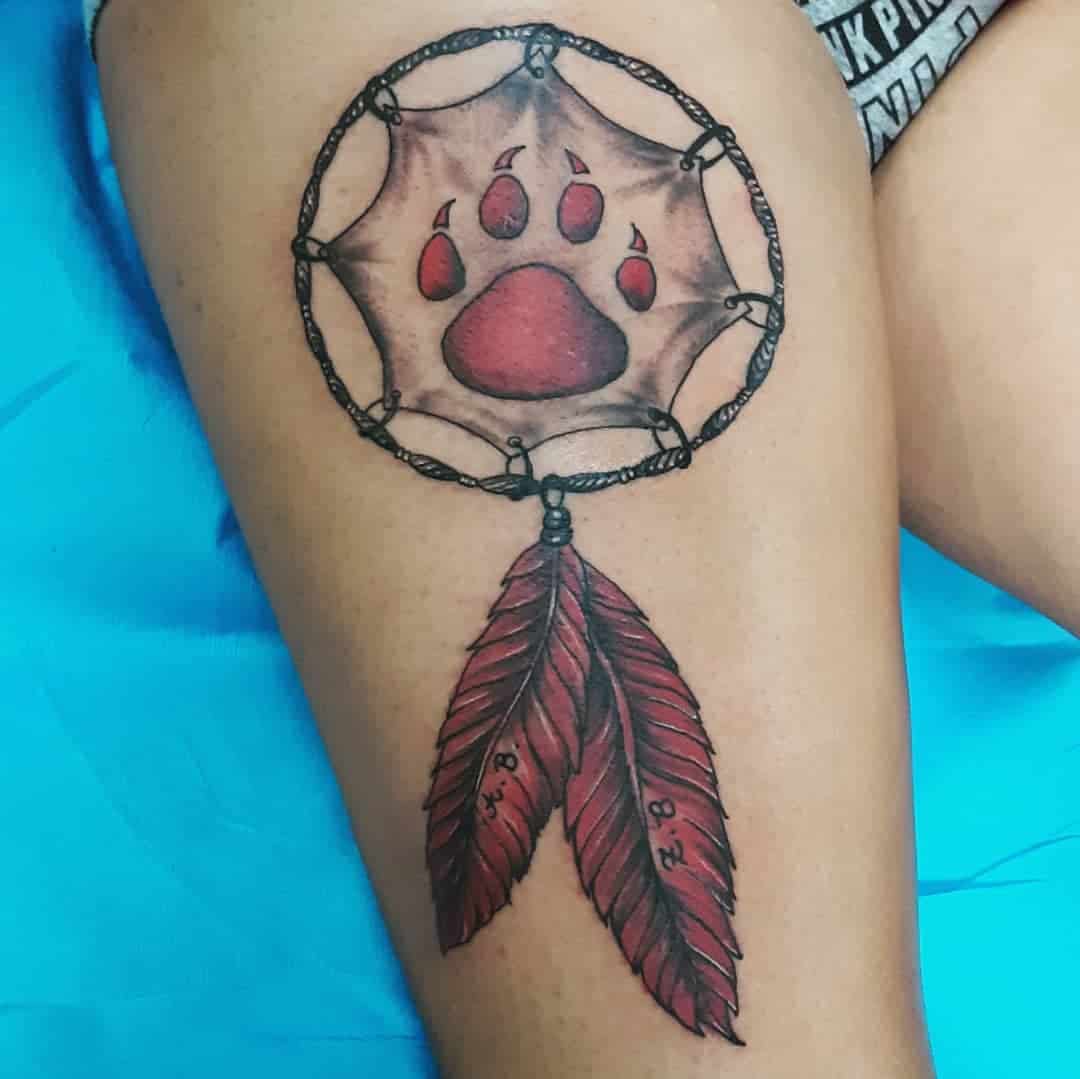 Tattoo of a dreamcatcher with a bear paw center and two hanging feathers on a person's thigh
