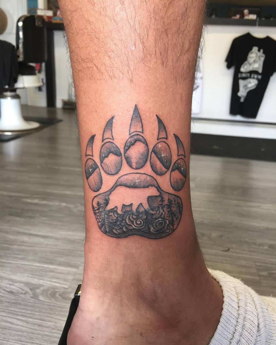 Bear paw tattoo with smaller bear and forest scene inside on person's ankle
