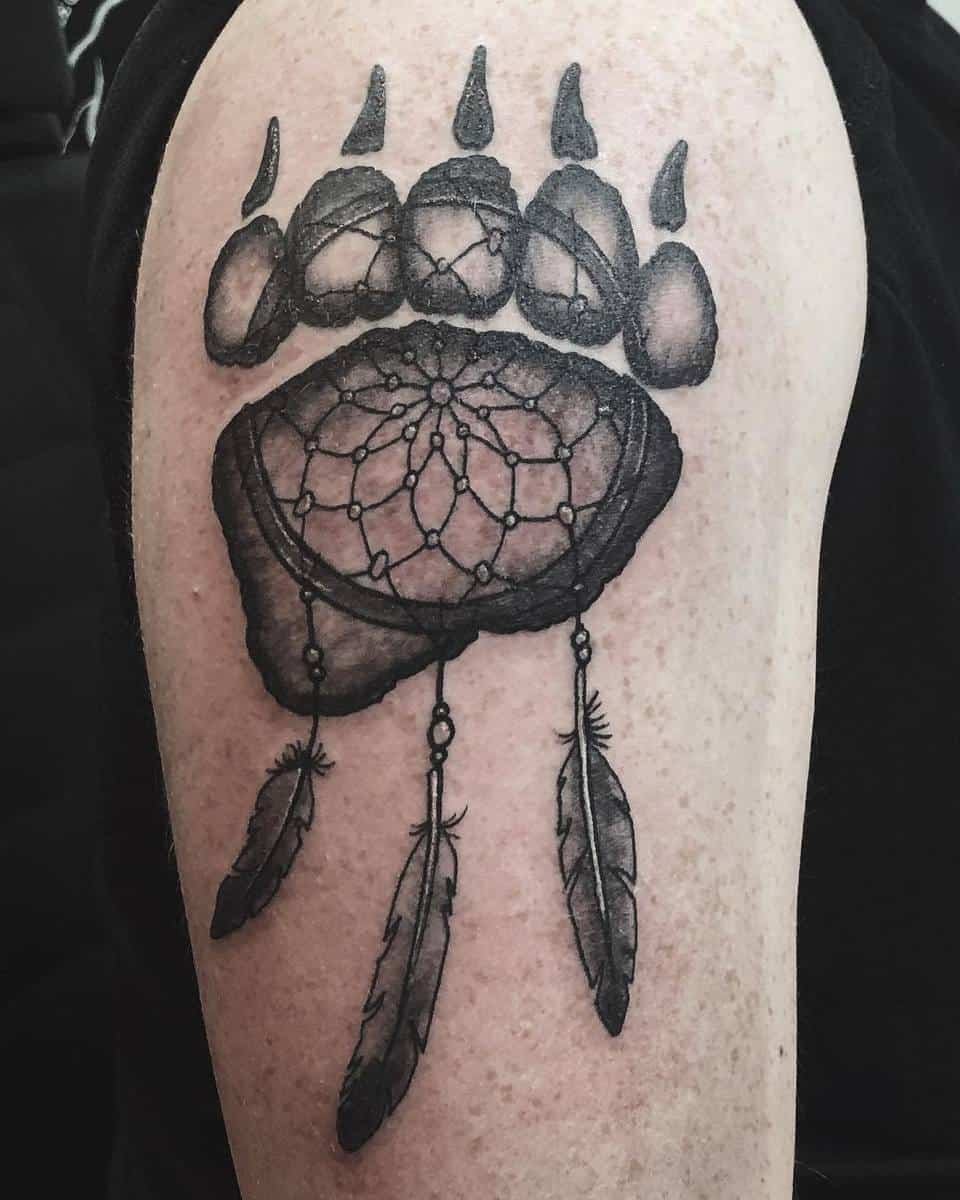 Tattoo of a bear paw with an intricate dreamcatcher design featuring feathers gracefully hanging from it on an upper arm