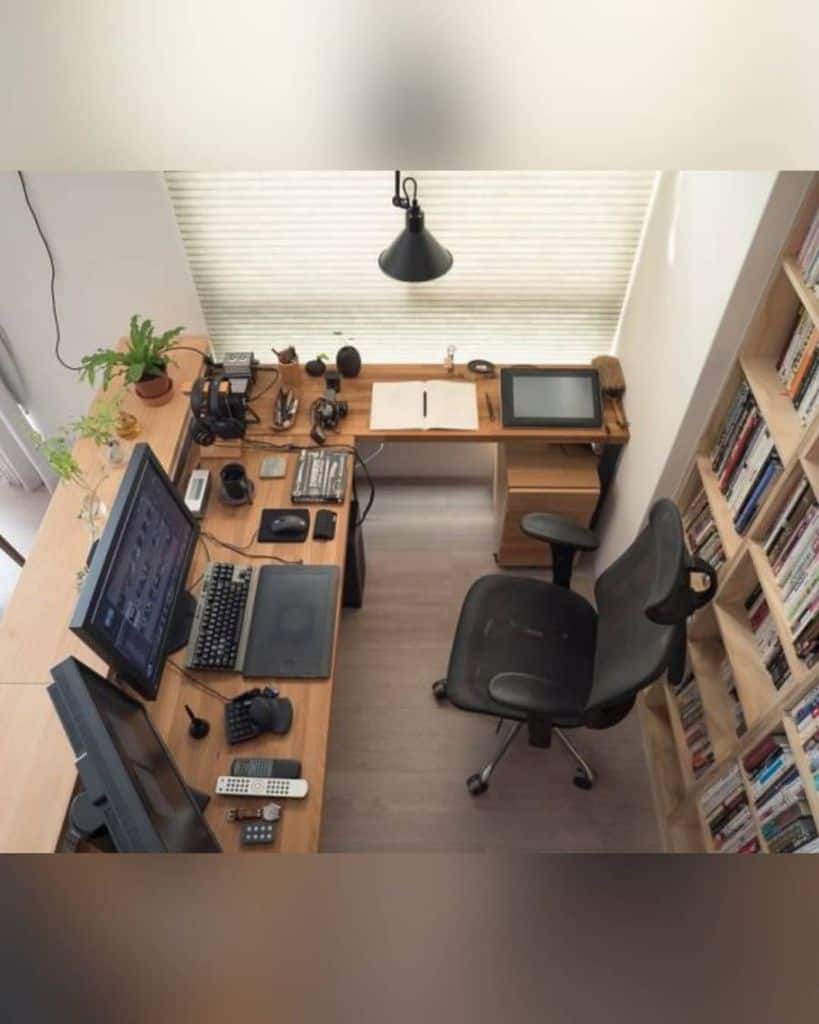 A cluttered home office with multiple screens, gadgets, a chair, bookshelves, and a window with blinds