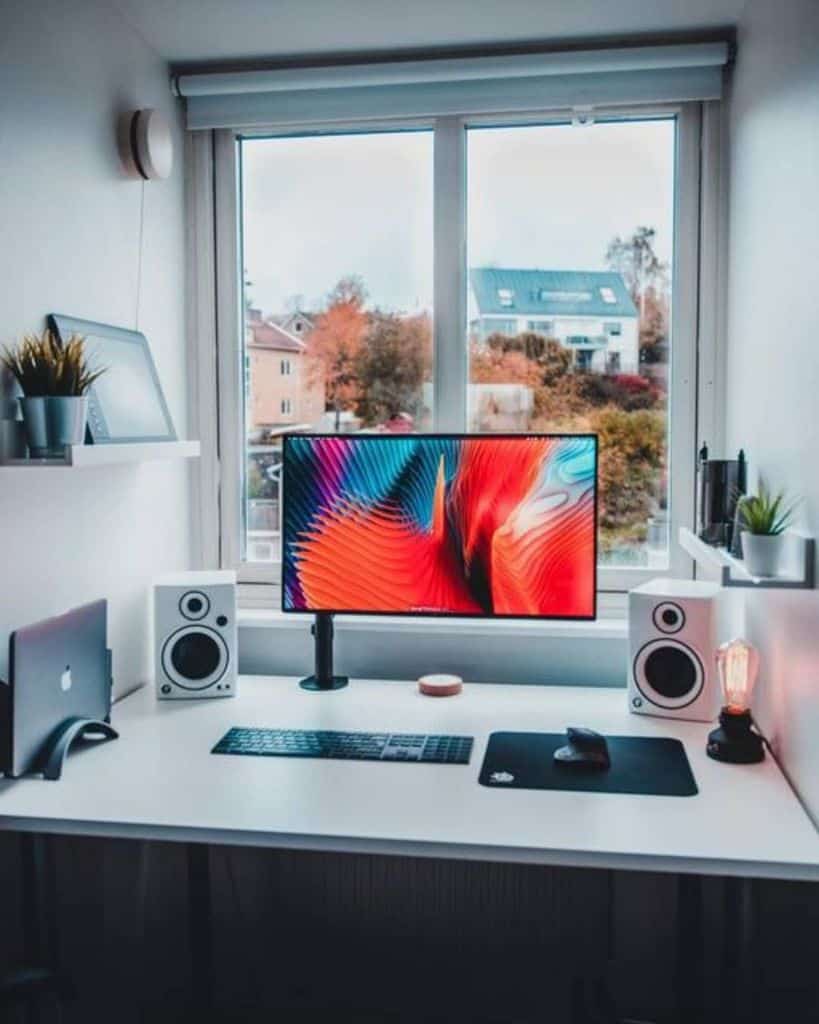 Minimalist desk setup with a monitor, speakers, and a laptop near a window; shelves with plants on the sides