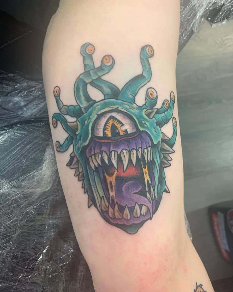 Bright and bold Beholder tattoo featuring a teal body, glowing orange eyes, sharp teeth, and a dynamic purple tongue