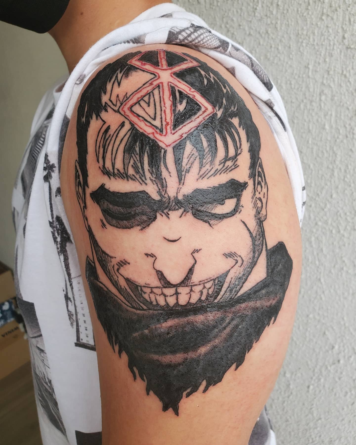 Tattoo of Guts from Berserk on the upper arm, featuring his intense grin and the Brand of Sacrifice prominently placed on his forehead