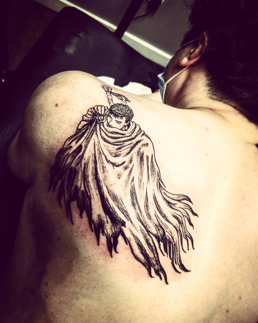 Tattoo of Guts from Berserk on the upper back, depicted in a dramatic pose with his cloak flowing and sword resting on his shoulder