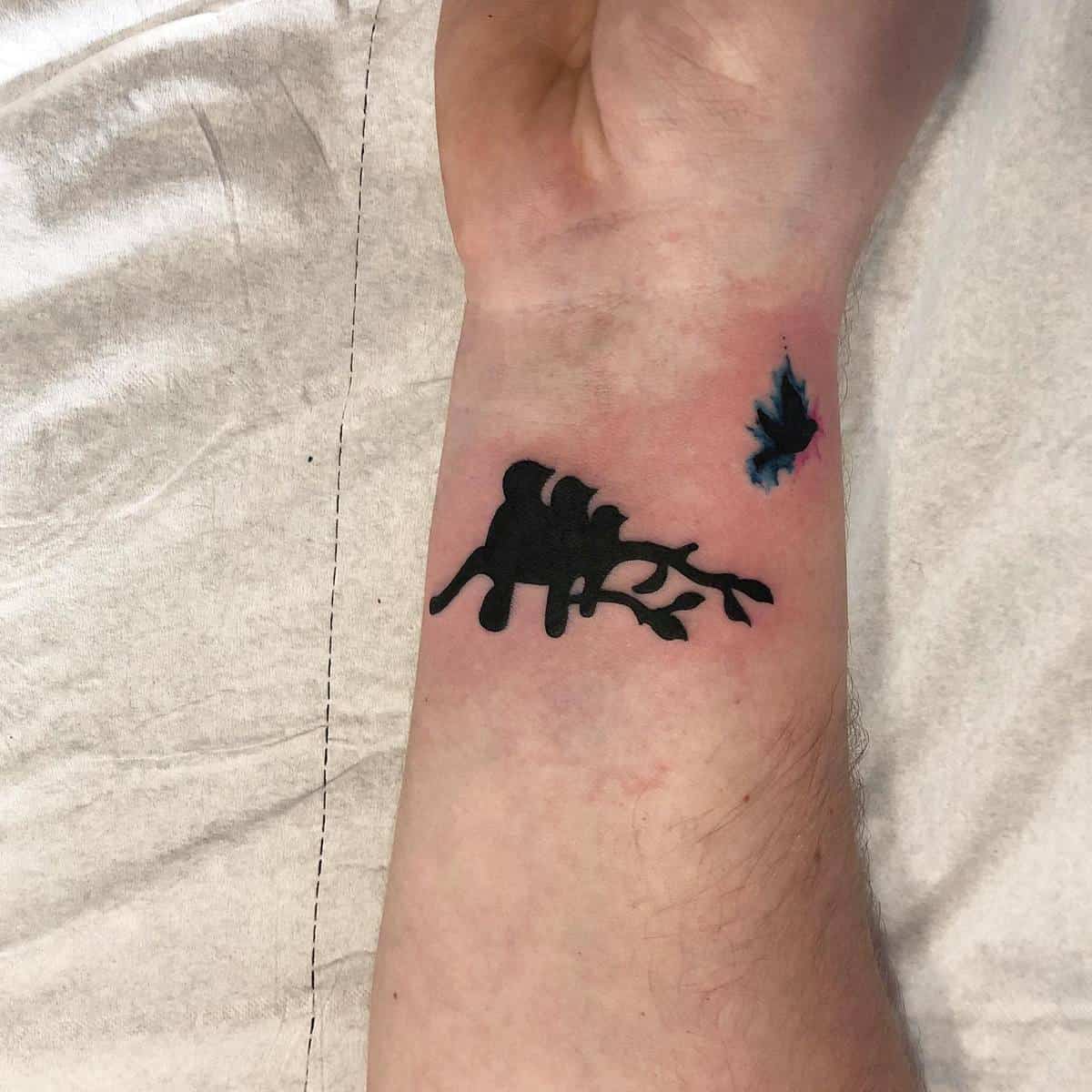 Forearm tattoo featuring a black, abstract tree design and a small, blue watercolor bird