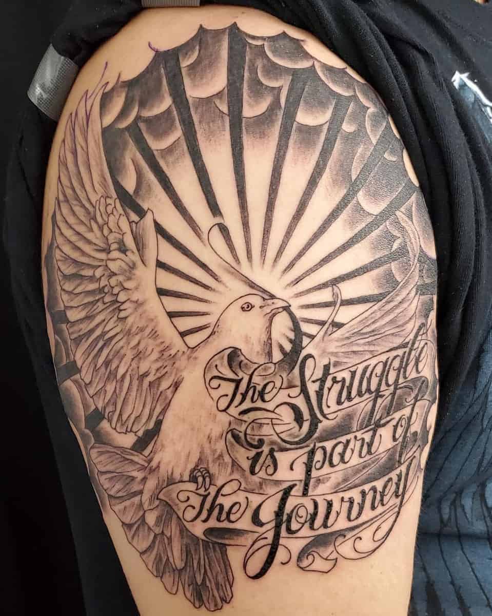 Tattoo on an upper arm depicting a bird with spread wings, rays, and the text "The struggle is part of the journey