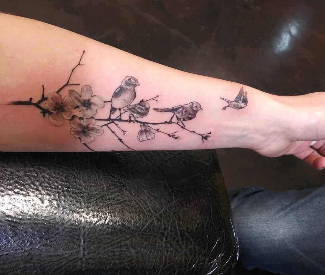 Forearm tattoo of birds with one bird flying away on a branch with flowers inked in black and white 
