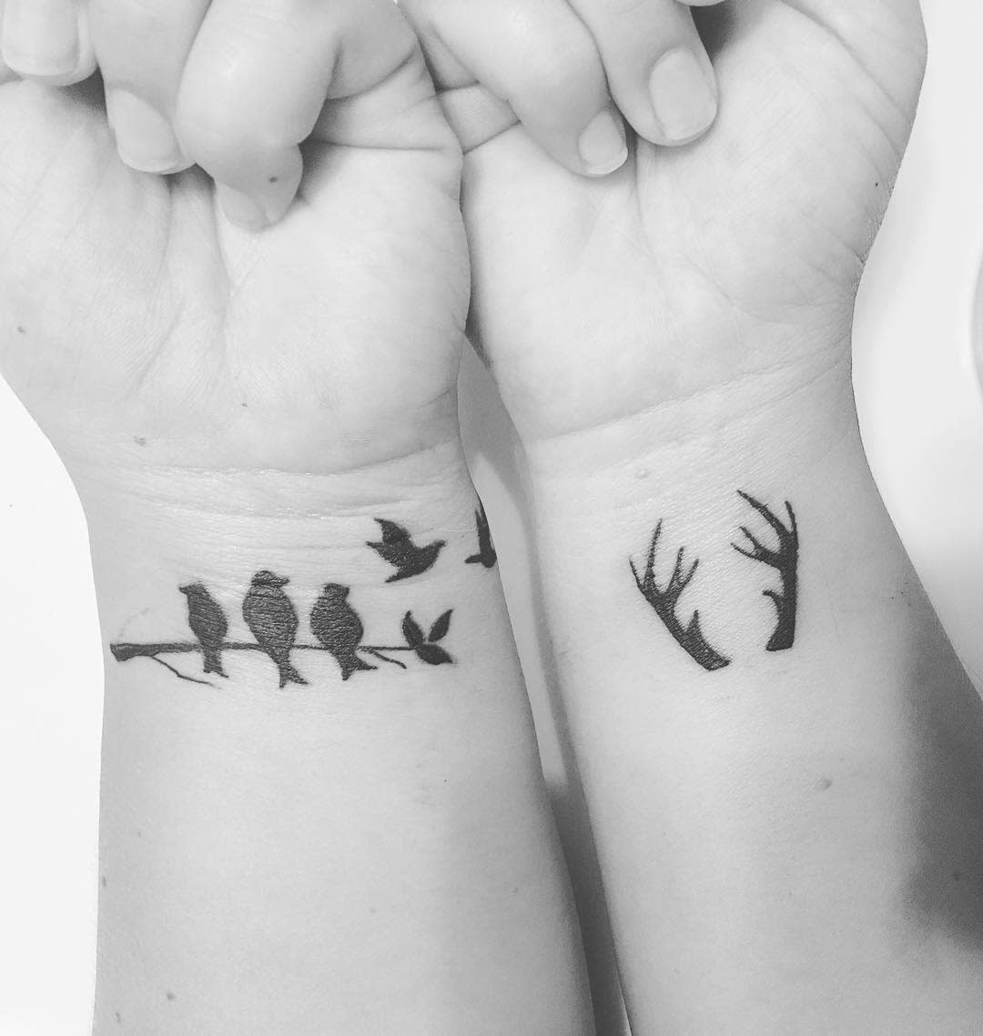 Two wrists with tattoos: the left wrist has birds on a branch, the right wrist has antlers, with hands clasped together