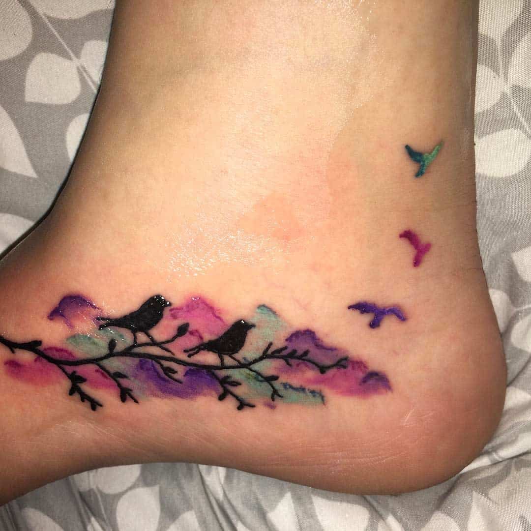 A tattoo on an ankle shows birds on a branch with colorful watercolor effects and three flying birds nearby