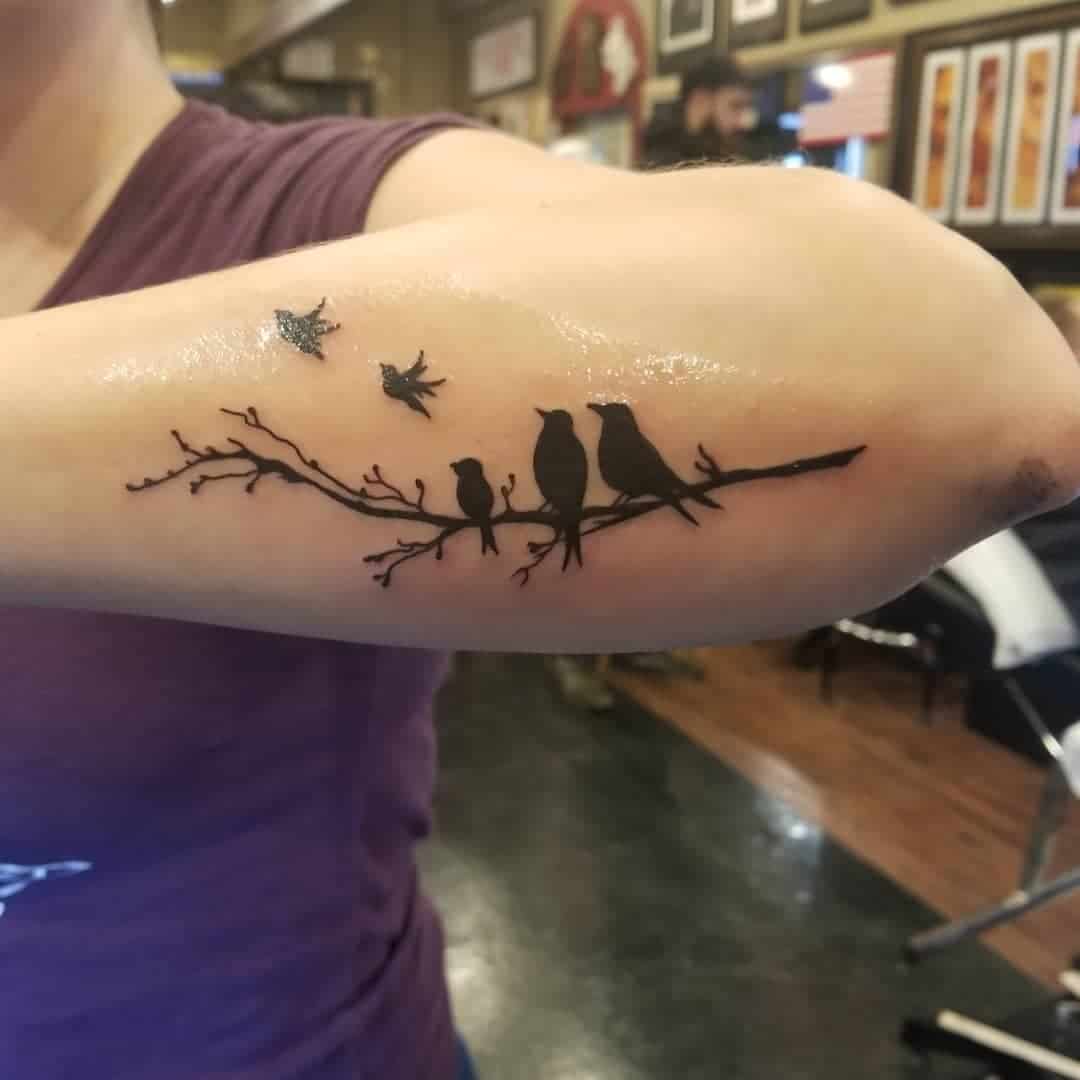 Tattoo of birds perched on a branch and two birds flying away on an upper arm
