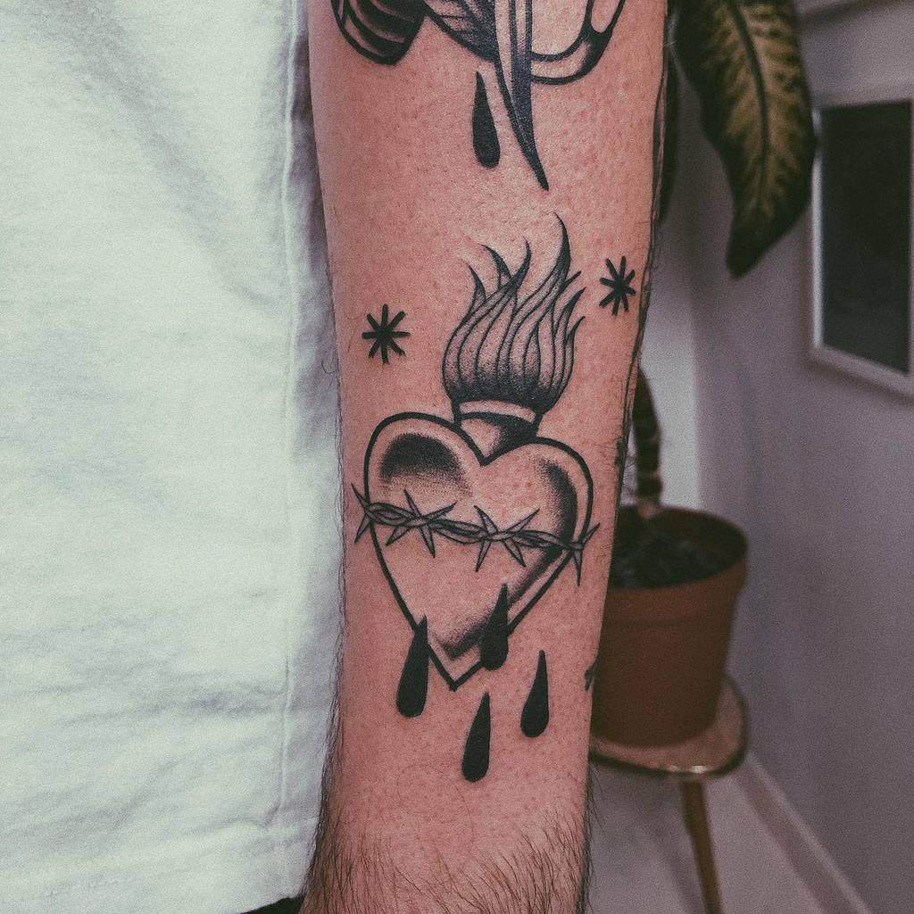 Tattoo of a flaming, bleeding heart with barbed wire and four drops, positioned on a person's forearm