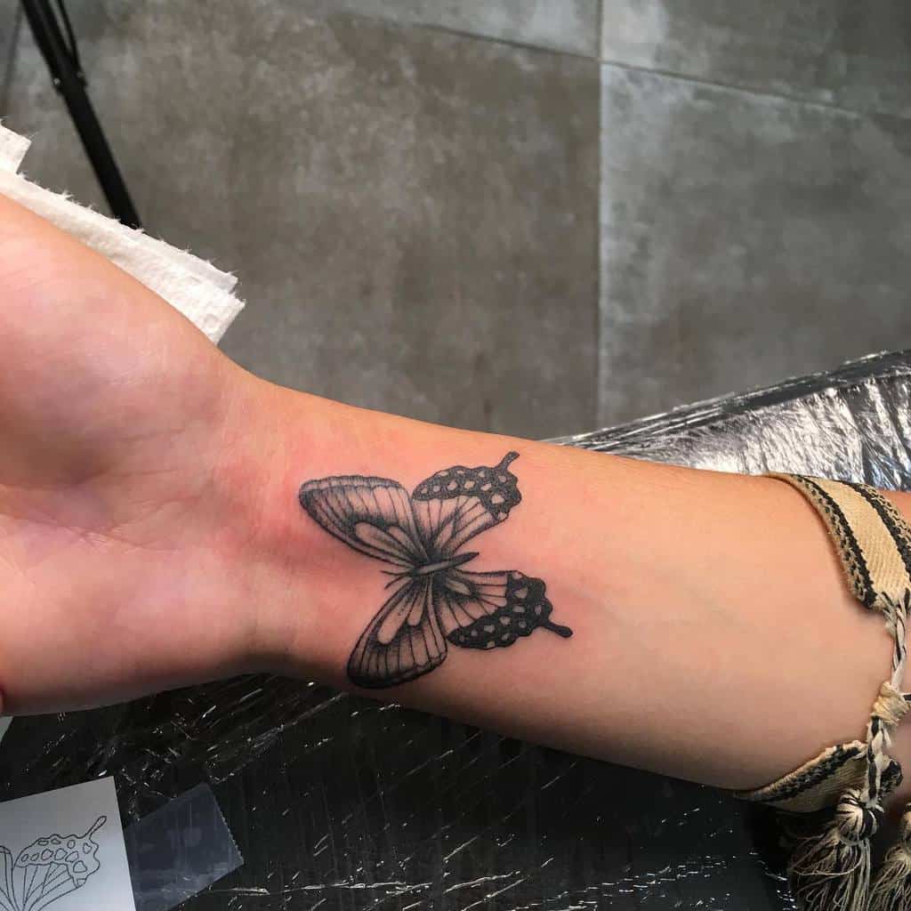 Wrist tattoo of a black butterfly with outlined, partially filled wings on a person's wrist