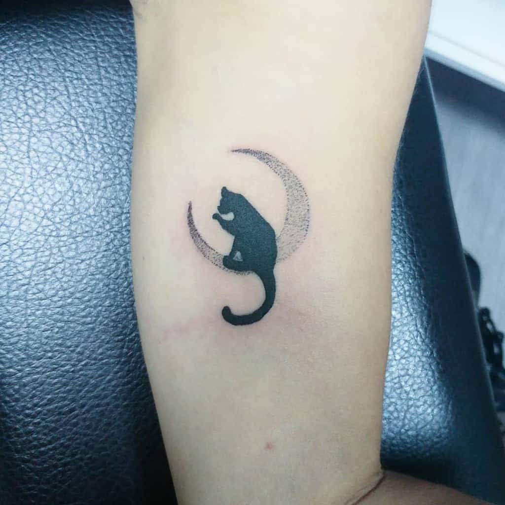 Small black cat and crescent moon tattoo on inner arm, showcasing sleek feline elegance