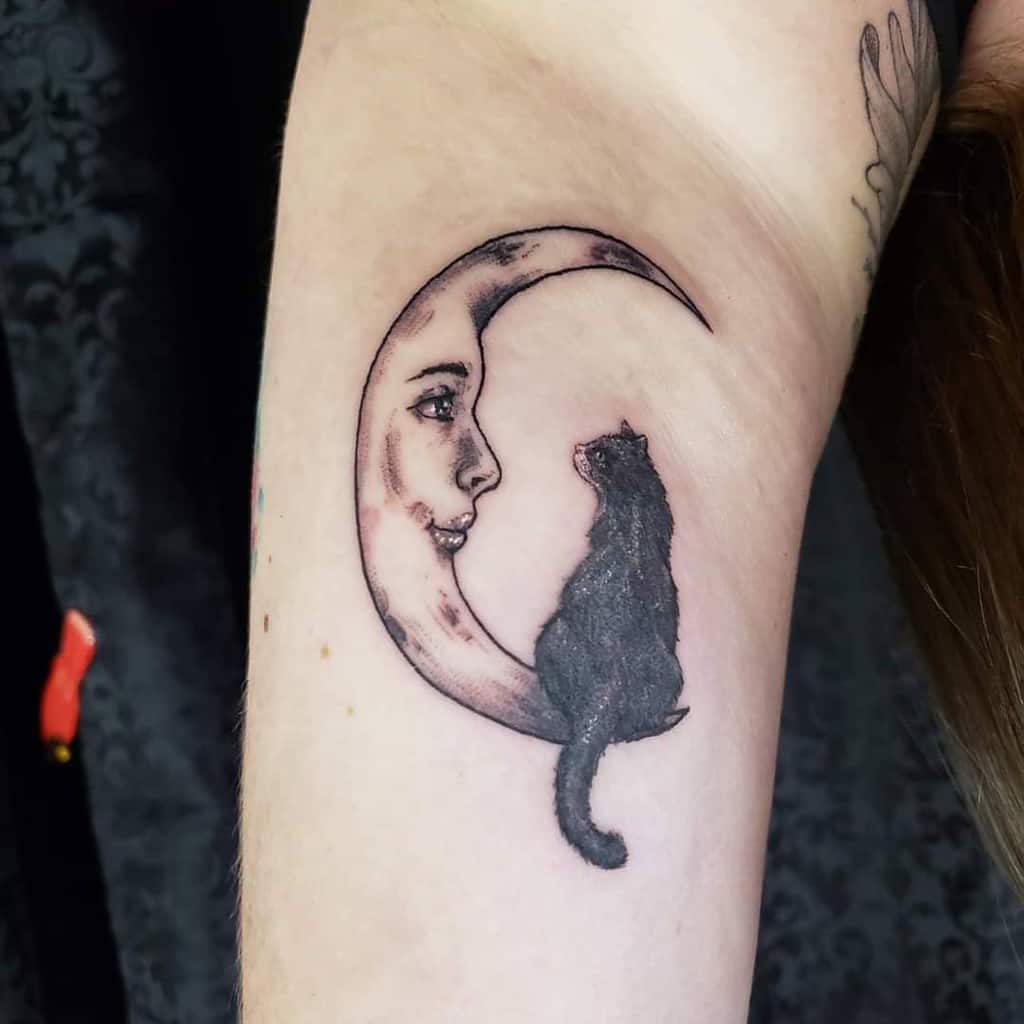 A tattoo of a moon with a face, adorned by a black cat gracefully perched on its tip, elegantly inked onto a person's arm