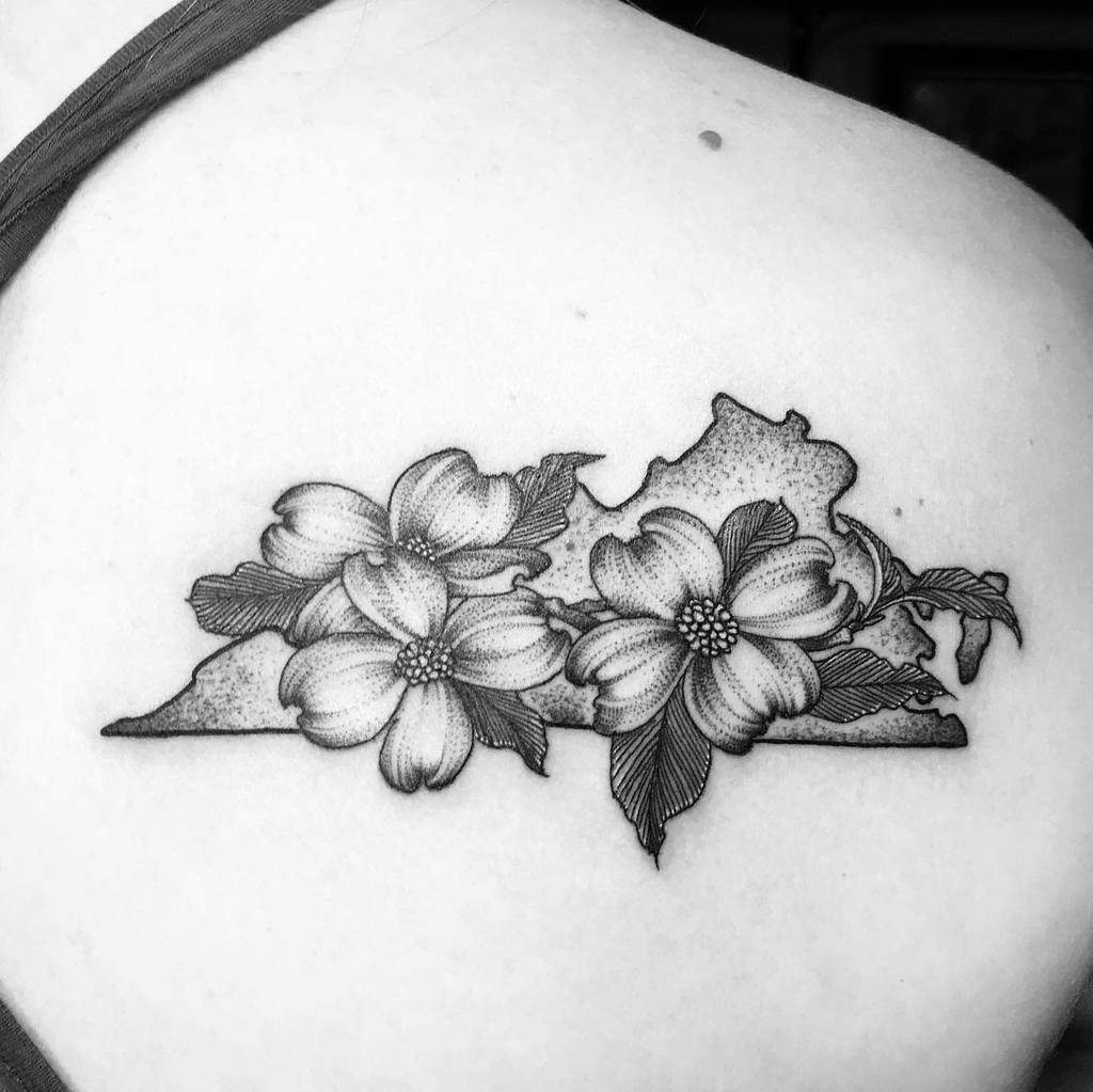A black and white tattoo of two dogwood flowers elegantly blooms over a state outline on a person's shoulder
