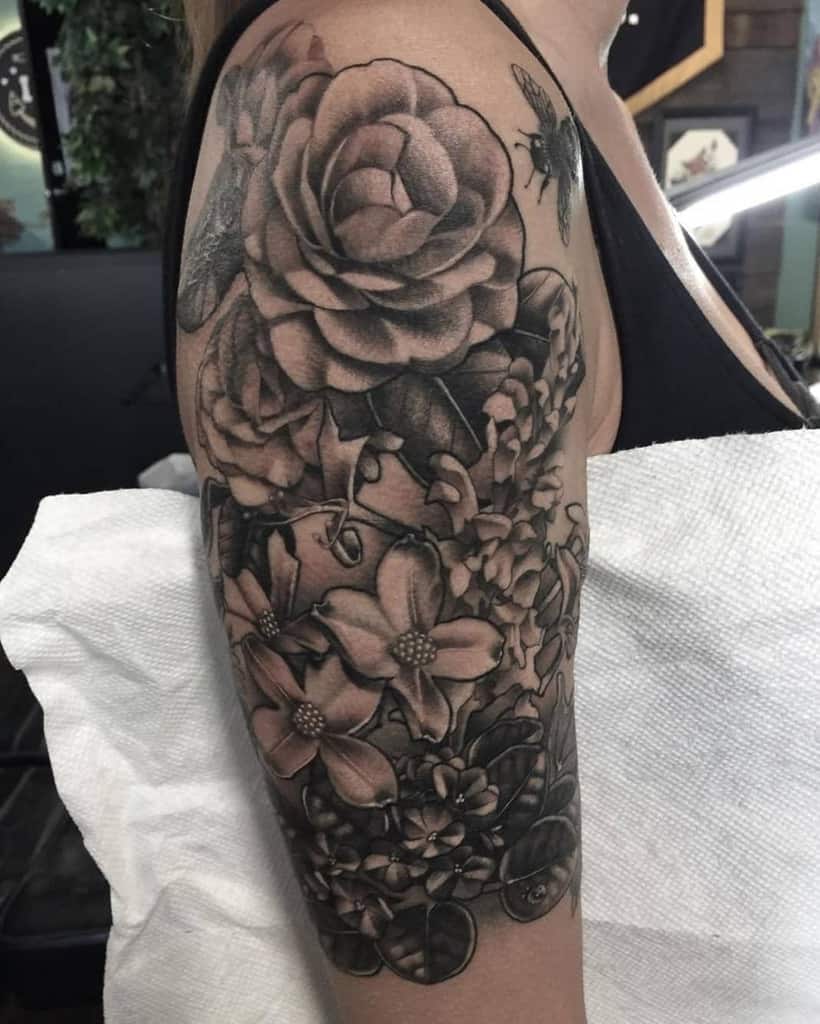An arm adorned with a detailed black and gray tattoo featuring intricate roses, delicate flowers, leaves, a bee, and elegant dogwood blossoms