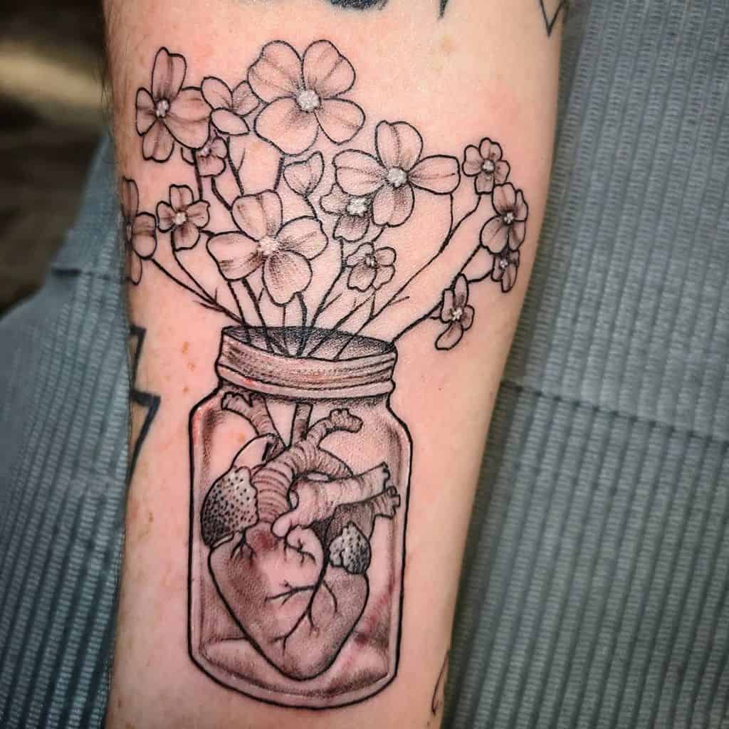 Unique black ink tattoo of a human heart placed inside a jar with dogwood flowers growing out of it, placed on the forearm