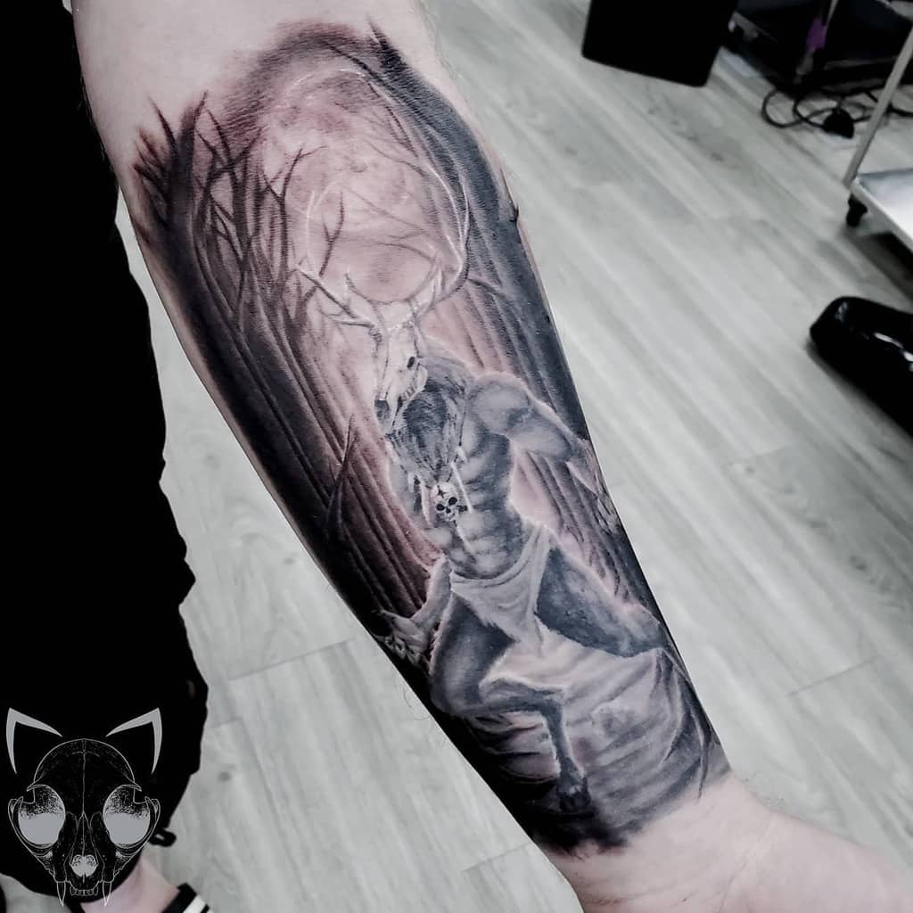 Dark forearm tattoo featuring a mythical stag-headed figure in a forest illuminated by a glowing moon