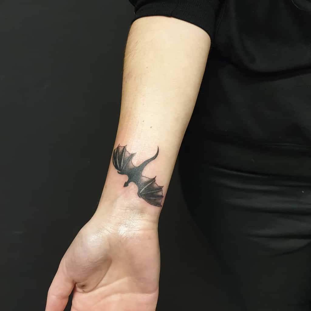 Small black dragon tattoo on the inner wrist, inspired by Game of Thrones, with outstretched wings and minimal detail