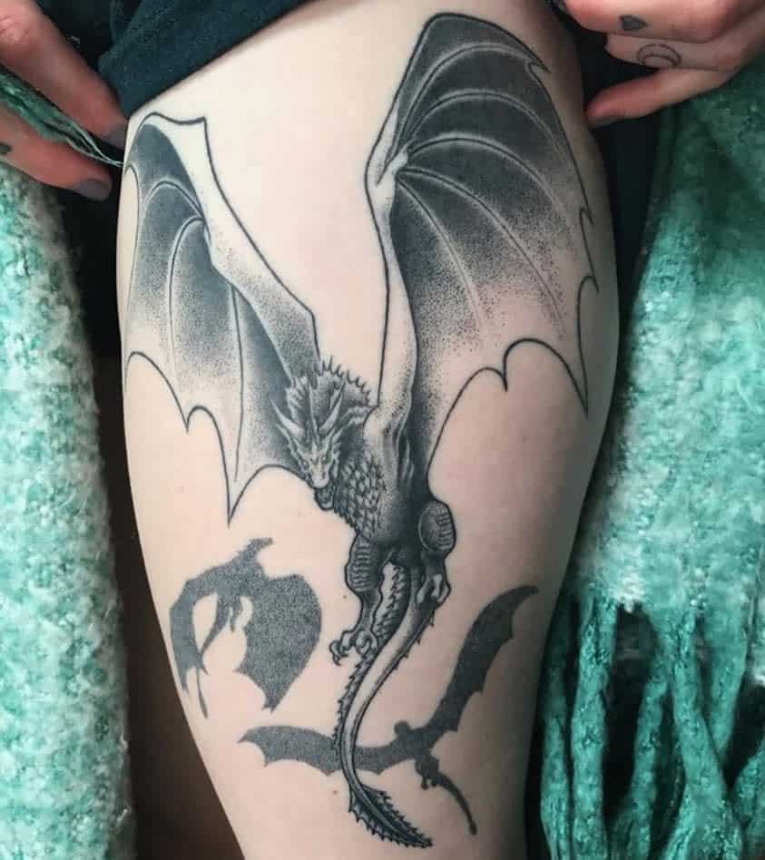 Detailed thigh tattoo of a dragon in flight with shaded wings, inspired by Game of Thrones, accompanied by smaller dragon silhouettes