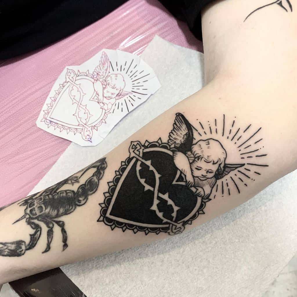 Detailed tattoo of a cherub clutching a black heart wrapped in thorns, surrounded by radiant light and decorative border