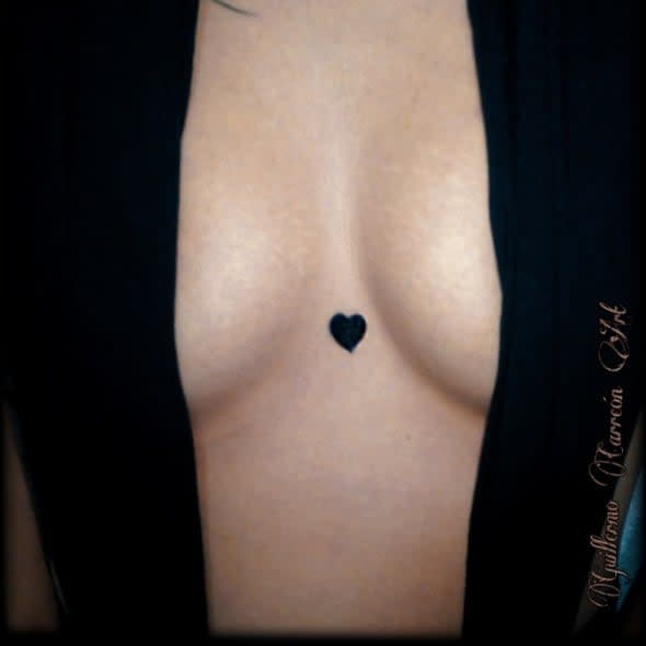 Minimalistic black heart tattoo centered on the sternum between the breasts, offering a bold and intimate statement