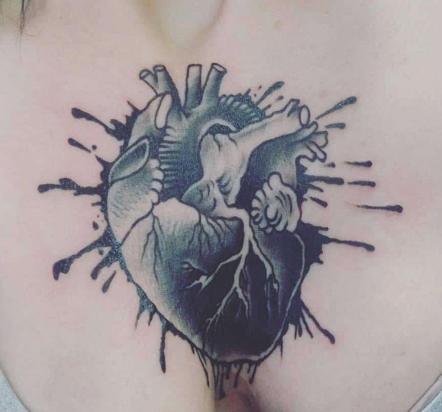 Tattoo of an anatomical black heart with ink splatters on skin, the design is detailed and shaded