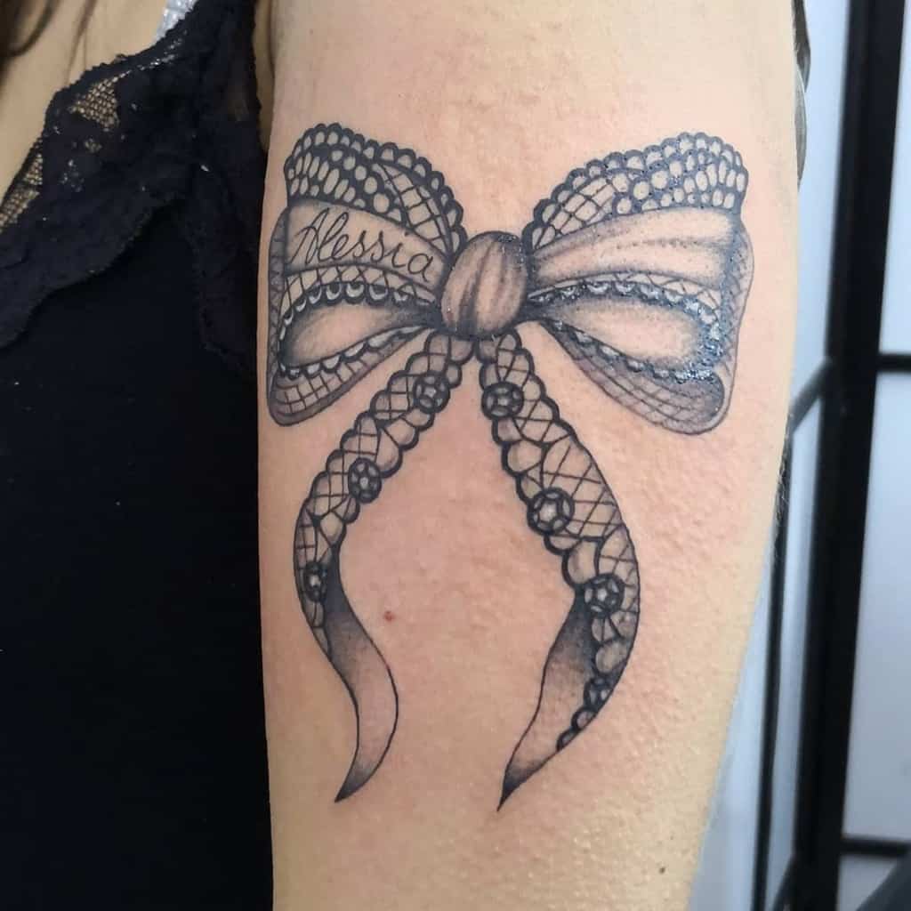 Delicate black and grey lace bow tattoo on the arm, featuring intricate details, beaded edges, and a name elegantly scripted on the ribbon.