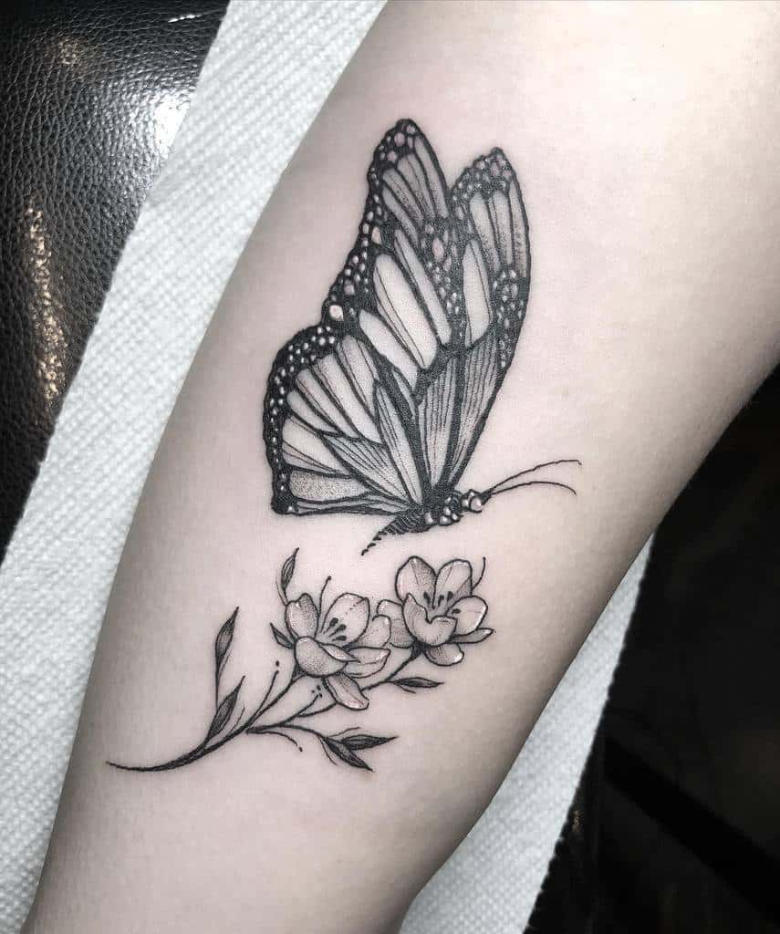A striking black and white tattoo of a monarch butterfly flutters above delicate small flowers on a person's forearm