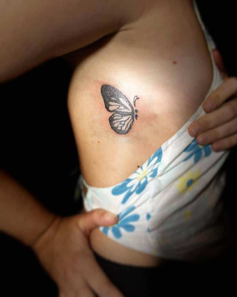 A small black semicolon butterfly tattoo on the ribcage, symbolizing resilience, hope, and mental health awareness