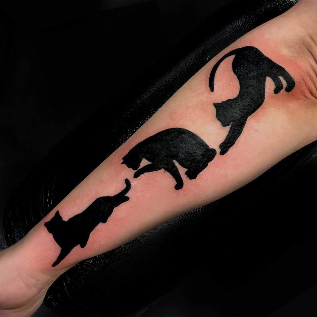 Three black cat silhouettes tattooed on a forearm, each in a different playful pose