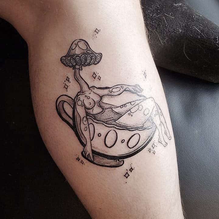 Tattoo of a relaxed figure lounging in a teacup with a mushroom umbrella, surrounded by small star-like shapes