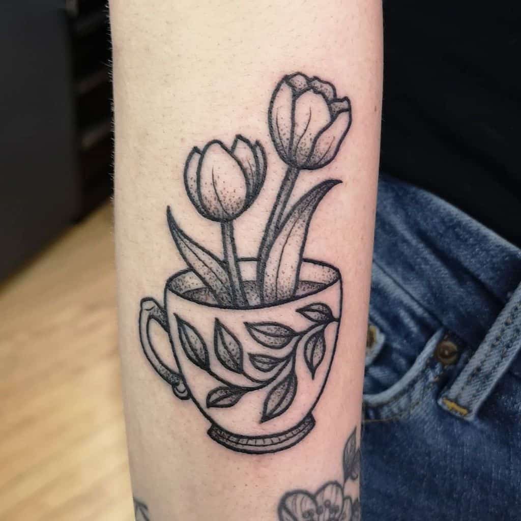 Black and white tattoo of a teacup with tulips growing from it, featuring delicate floral details in an elegant design