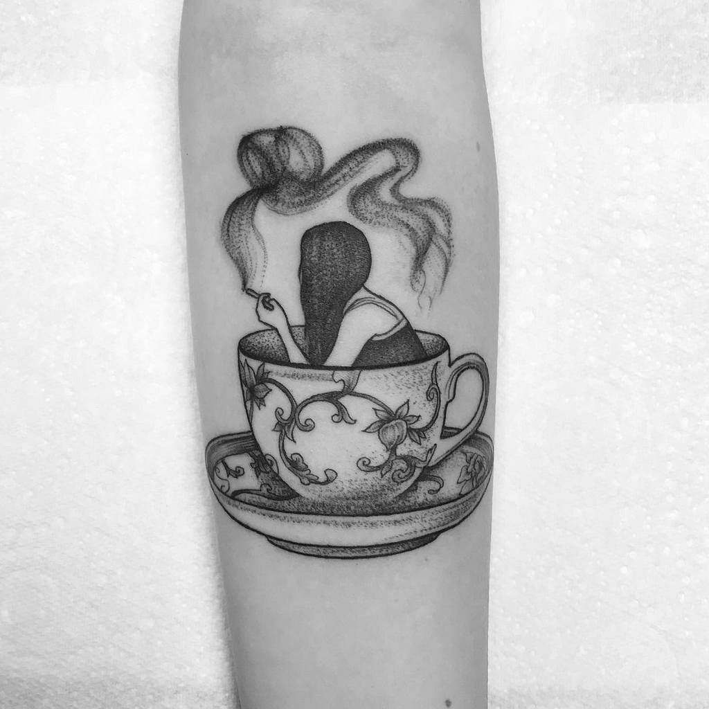 Black and white tattoo of a woman sitting inside an ornate teacup, smoking a cigarette, with swirling smoke rising above her