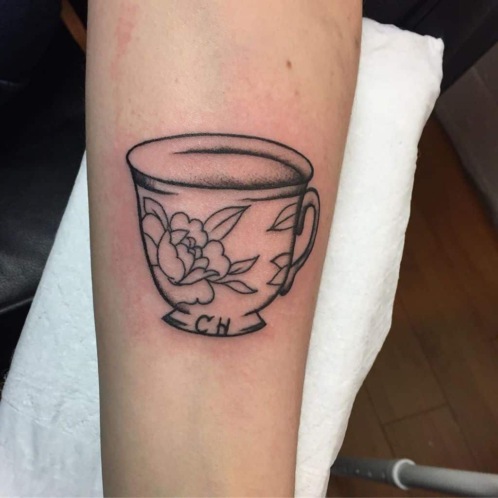 Black and white tattoo of a teacup with floral patterns and the initials "CH" on the base, featuring a shaded liquid effect on top