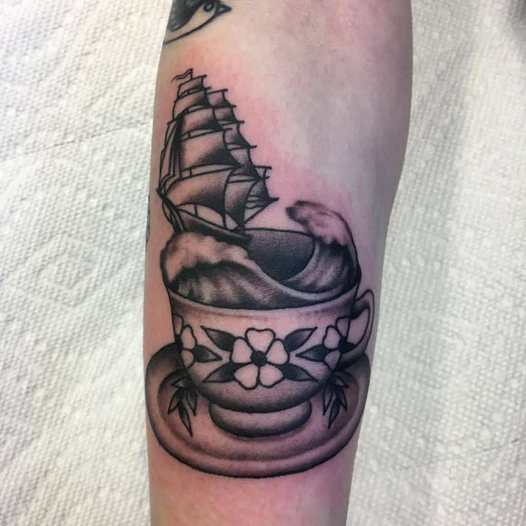 Black and white tattoo of a teacup with waves inside, featuring a sailing ship emerging from the cup, creating a surreal nautical theme