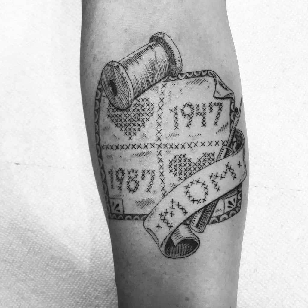 Intricate black and grey tattoo of a cross-stitched fabric with dates '1947', '1987', hearts, and a 'MOM' ribbon with thread