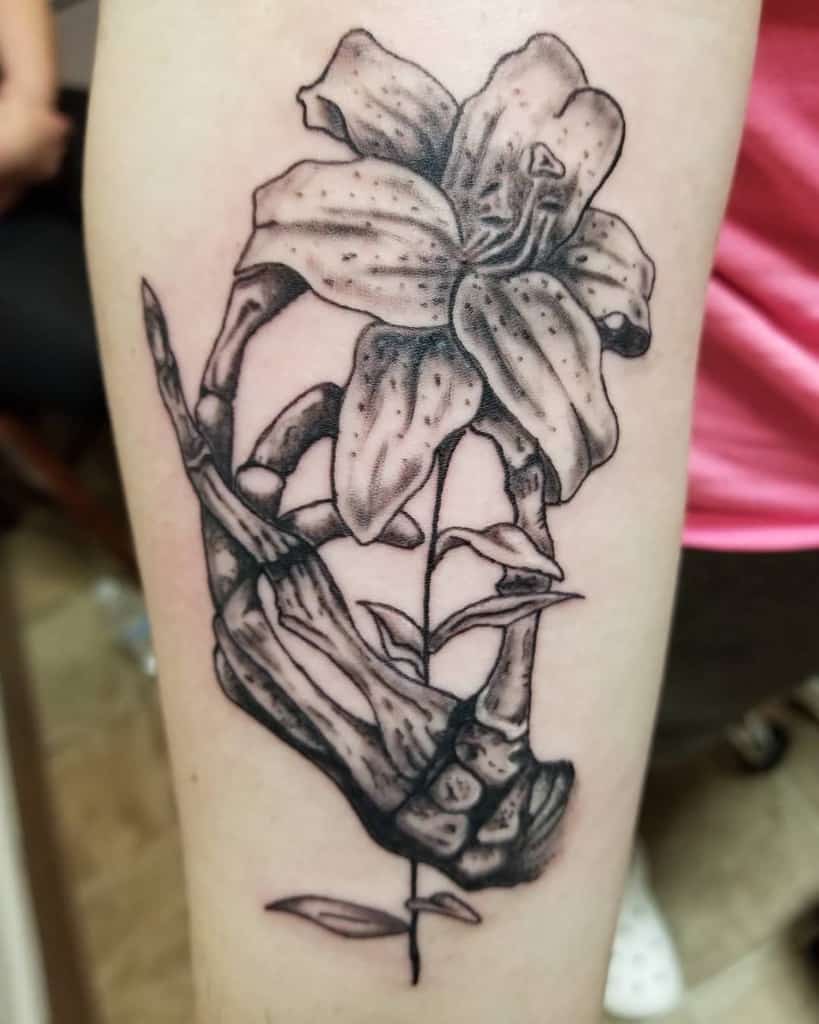 Bold black and grey tattoo of a skeletal hand holding a blooming lily, symbolizing life, death, and the beauty of fleeting moments