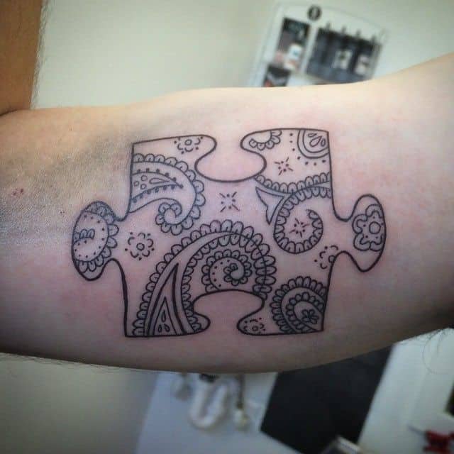 Black and gray bicep tattoo of a puzzle piece with paisley designs inside.