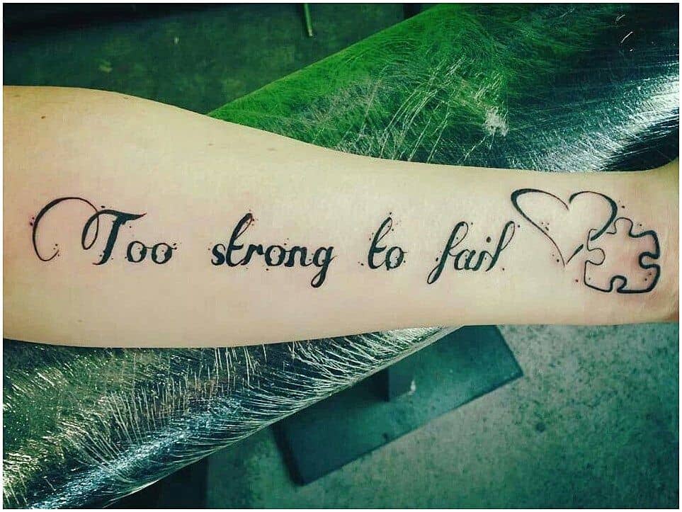 Black forearm tattoo “Too strong to fail” with a heart and puzzle piece.