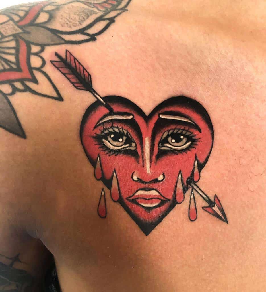 A bleeding heart tattoo with a face, tears, and an arrow piercing through it on a person's shoulder