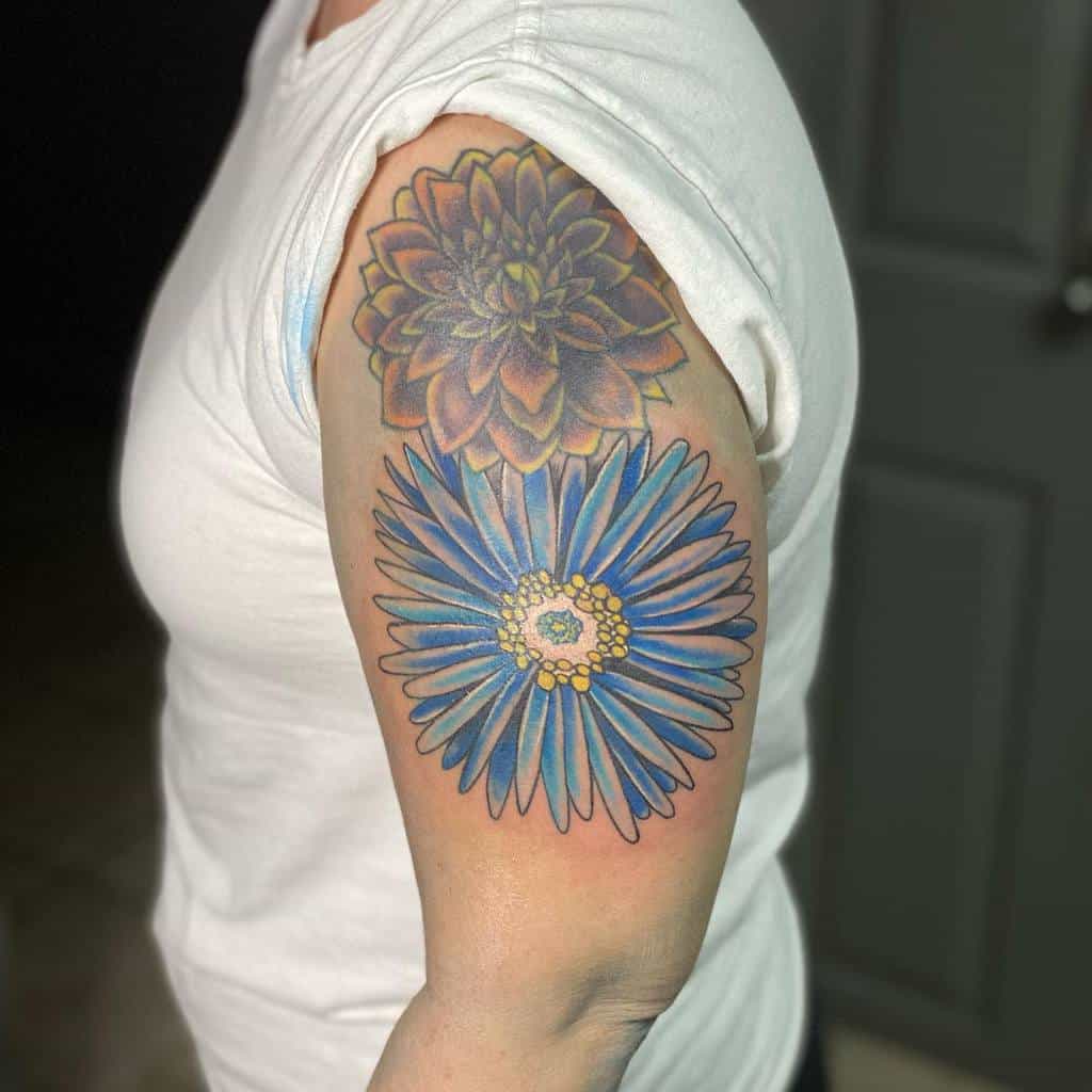 A colorful upper arm tattoo featuring a detailed blue aster flower with yellow accents