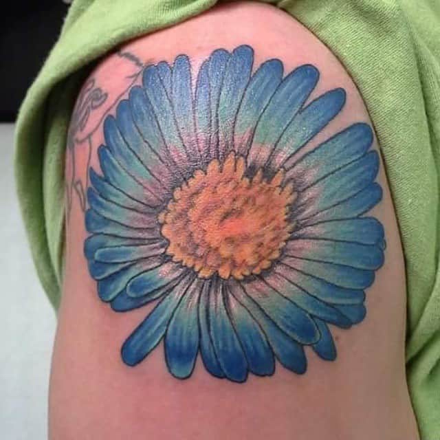 A vibrant blue and orange aster flower tattoo on an upper arm