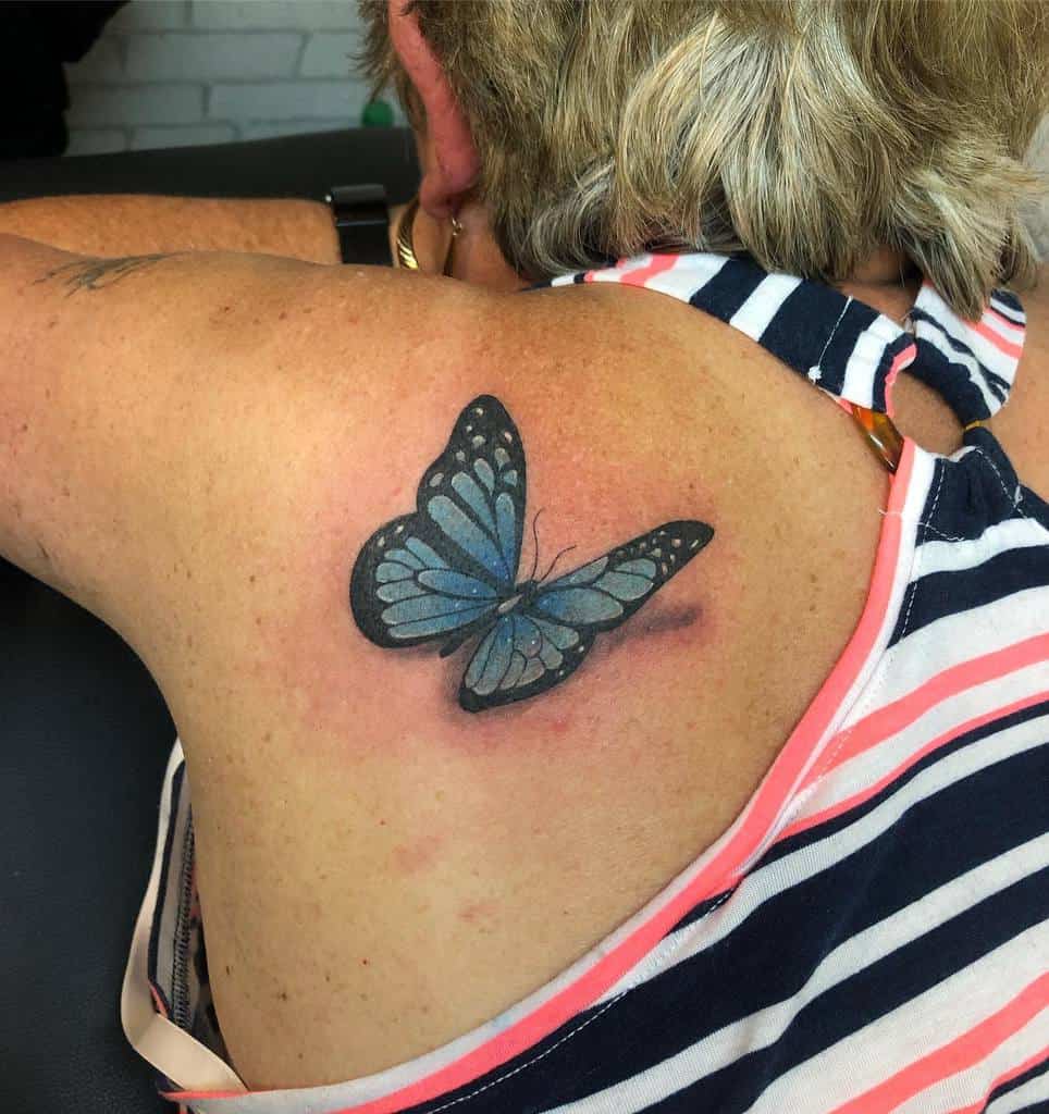 Realistic blue butterfly tattoo on upper back, designed with a 3D effect, creating the illusion of the butterfly resting on the skin.