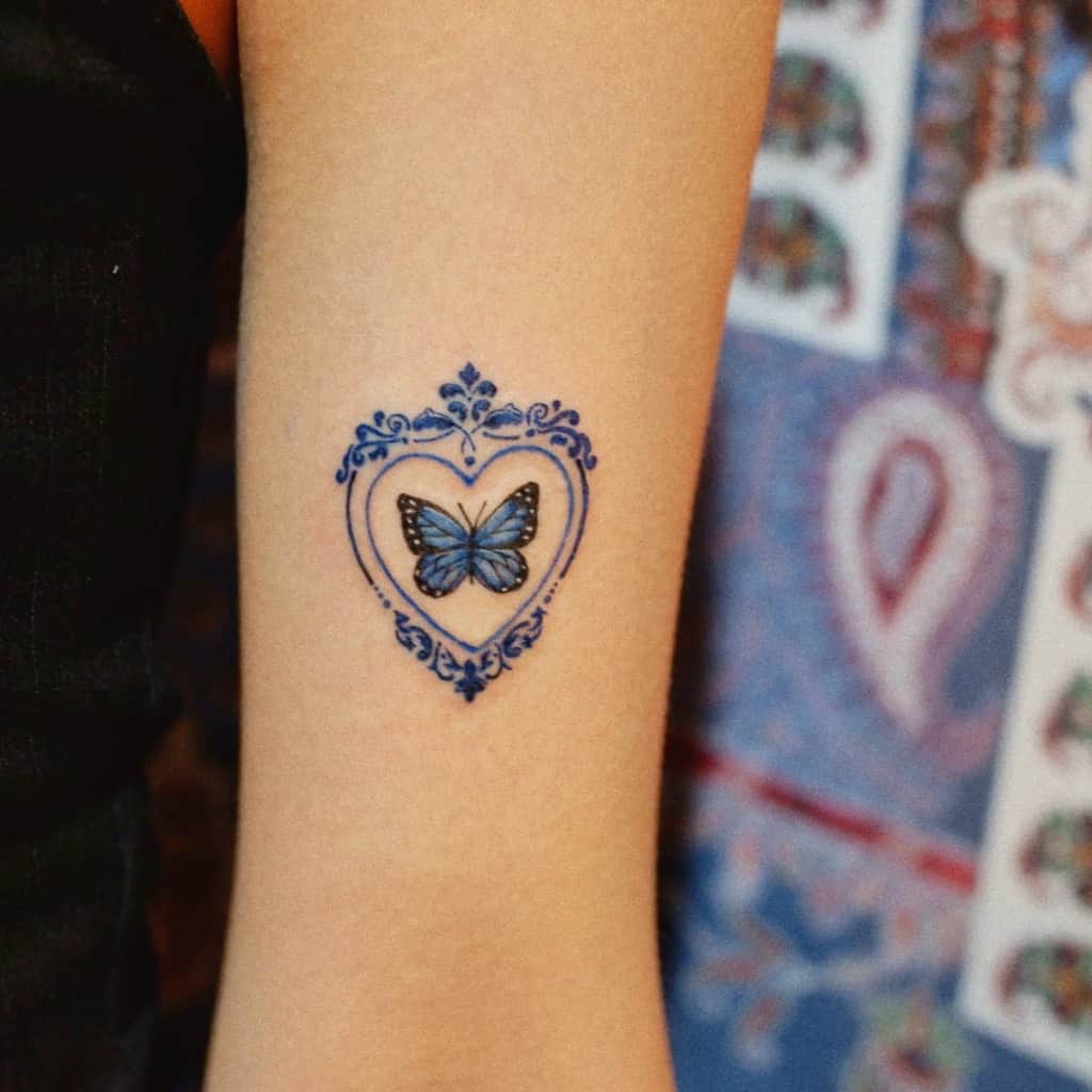 Delicate blue butterfly tattoo inside an ornate heart-shaped frame on the arm, featuring intricate decorative details.