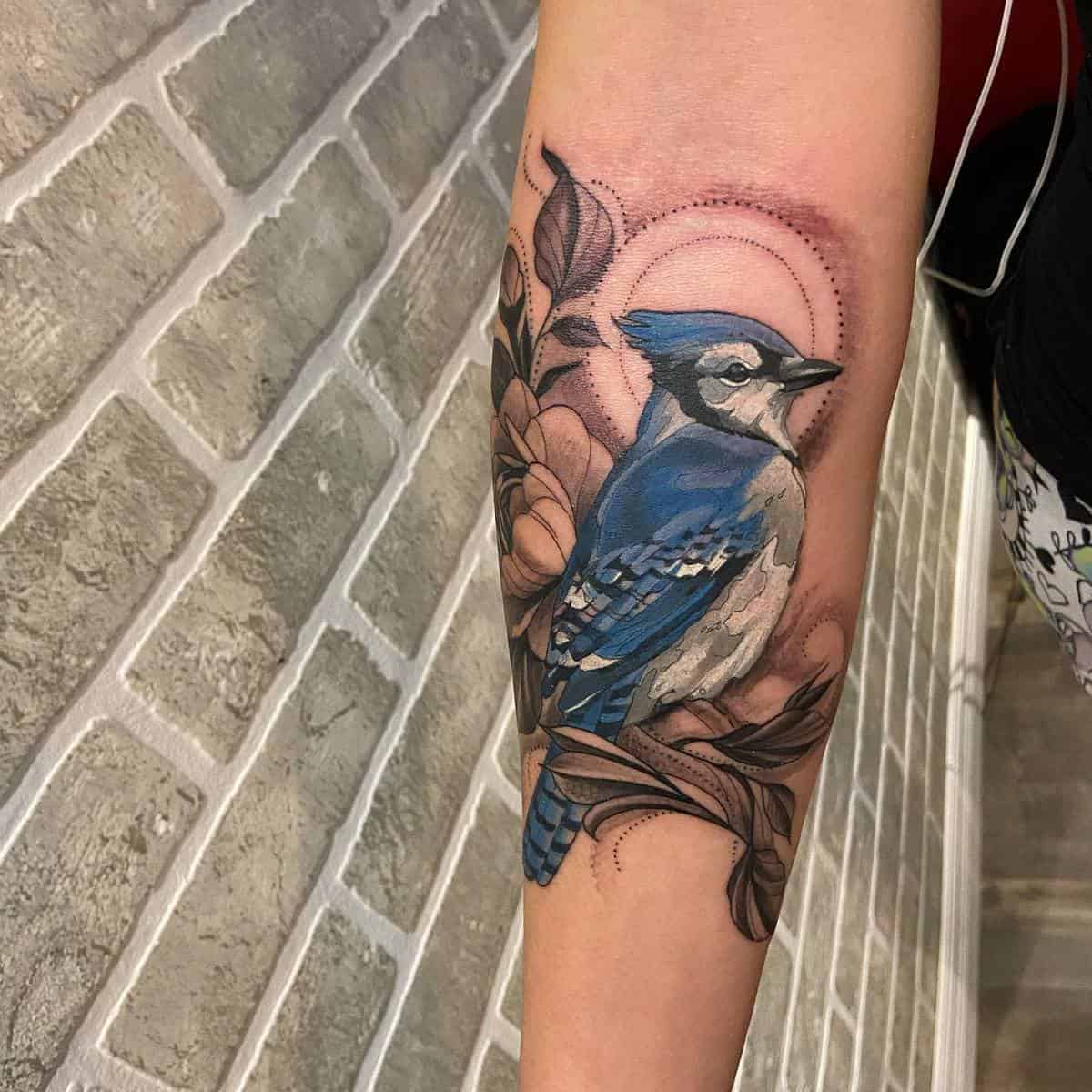 A forearm tattoo of a blue jay perched on a branch with flowers and geometric patterns